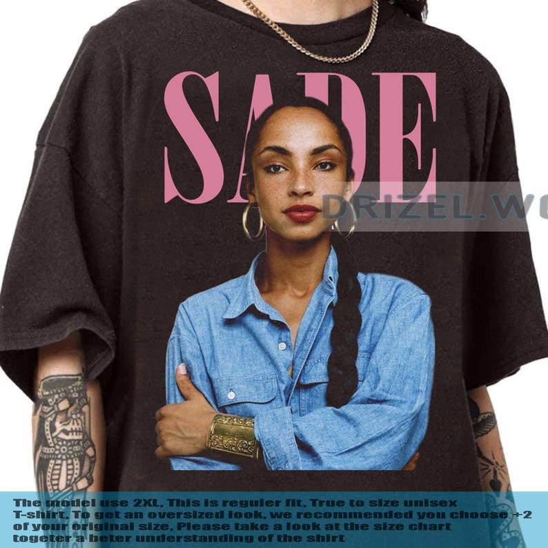 May include: Black unisex t-shirt with a portrait of Sade Adu and the name "SADE" in pink above. The model wears a denim shirt. The t-shirt is regular fit and true to size. The text at the bottom is blurry.