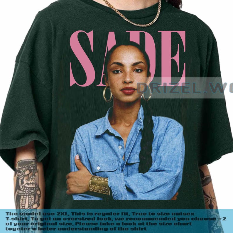 May include: Dark green unisex t-shirt with a portrait of Sade Adu and the name "SADE" in pink. The image shows a woman in a denim shirt and gold jewellery. The text at the bottom says the model is wearing 2XL.