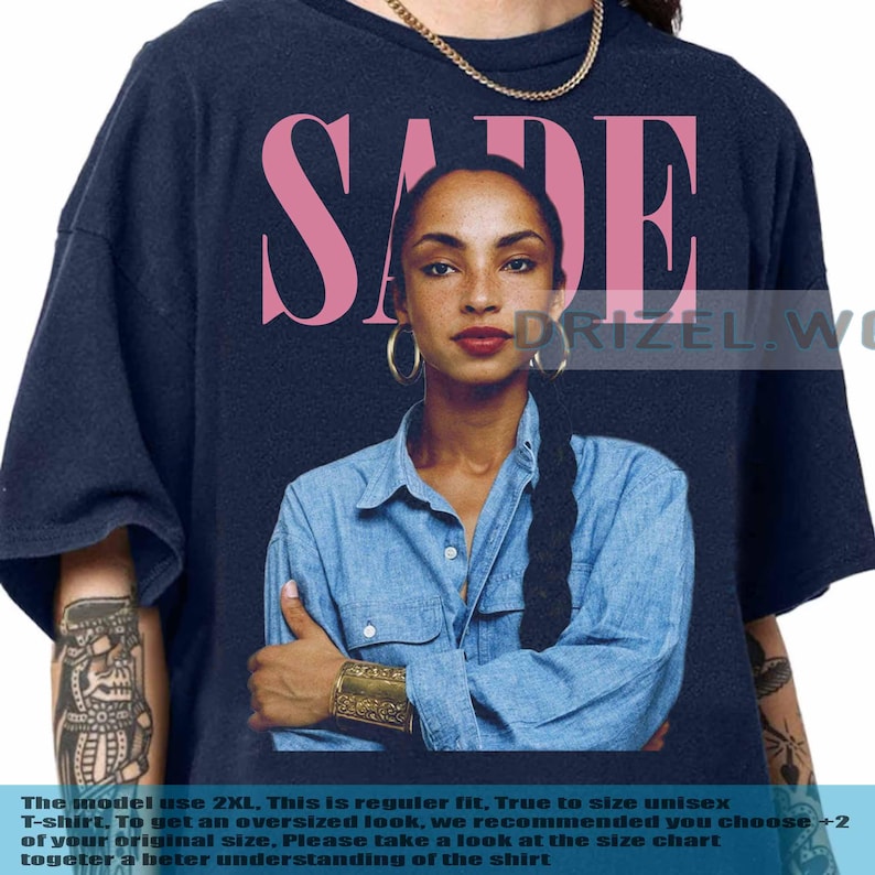 May include: Navy blue t-shirt with the pink word "SADE" above a portrait of a woman in a denim shirt and gold jewellery. The image includes the text "DRIZEL.WO" on the right. The model is wearing a gold chain.