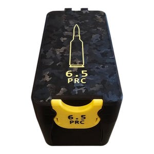 Könnte beinhalten: Eine schwarze Munitionskiste mit einem Tarnmuster auf dem Deckel. Der Deckel zeigt eine goldene Umrisszeichnung einer Kugel und den Text "6.5 PRC". Die Kiste hat einen gelben Verschluss mit dem gleichen Text.