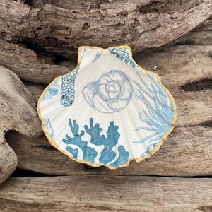 May include: A decorative seashell-shaped dish with a white interior featuring blue coral and seashell designs. The rim is edged with gold. The dish is resting on a background of weathered driftwood.