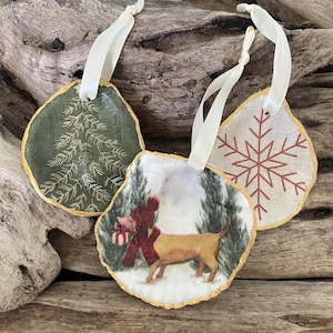 May include: Three Christmas tree decorations with cream ribbon hangers. One has a green background with a white tree design. Another features a dog wearing a hat and scarf in a winter scene. The third has a red snowflake design.
