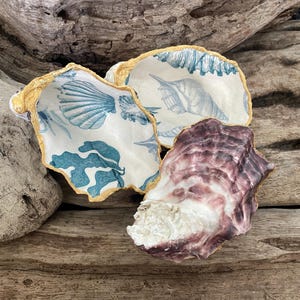 May include: Three decorative oyster shells. Two shells are painted white with blue seashell patterns and gold trim. The third shell is a natural purple and white colour. The shells are arranged on a weathered wooden surface.