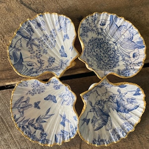 May include: Four seashell-shaped decorative dishes with blue floral and butterfly patterns on a white background. The edges are trimmed with gold. The dishes are arranged on a wooden surface.