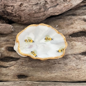 May include: A white oyster shell dish with a gold rim, decorated with four yellow and black bee illustrations. The shell is set on a weathered, brown wooden surface, creating a natural, rustic aesthetic.