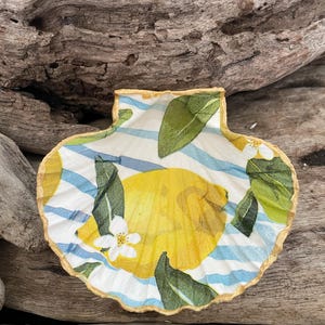 May include: A decorative scallop shell with a lemon and leaf design. The shell features a yellow lemon, green leaves, and white flowers against a backdrop of blue and white stripes. The shell's edge is trimmed with a gold colour.