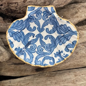 May include: A decorative seashell dish featuring a blue and white floral design. The shell has a gold-coloured rim and is set against a backdrop of weathered wood.
