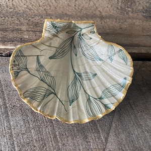 May include: A decorative seashell-shaped dish with a beige interior and a gold-coloured rim. The dish features a green leaf pattern. The seashell dish is a decorative item.