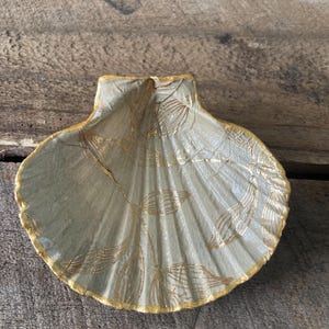 May include: A decorative seashell-shaped dish with a pale green base and gold leaf detailing. The rim is trimmed with gold paint. The dish is on a wooden surface.