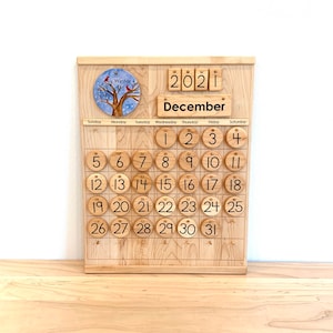 May include: A wooden perpetual calendar featuring a light-coloured wood grain finish. It displays the year 2021 and the month December. A circular disc shows a winter tree with red birds. Round wooden tiles numbered 1 through 31 are arranged.