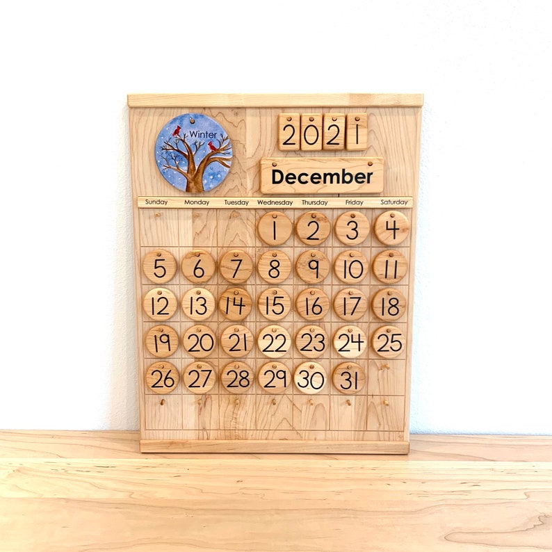 May include: A wooden perpetual calendar featuring a light-coloured wood grain finish. It displays the year 2021 and the month December. A circular disc shows a winter tree with red birds. Round wooden tiles numbered 1 through 31 are arranged.