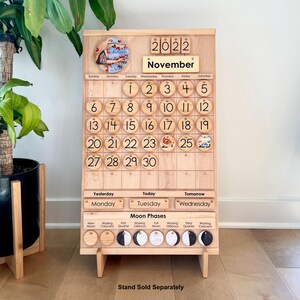 May include: A light-coloured wooden perpetual calendar board featuring the year 2022 and the month of November. It includes numbered circular wooden discs for dates, a section for days of the week, and a moon phases tracker with circular icons.