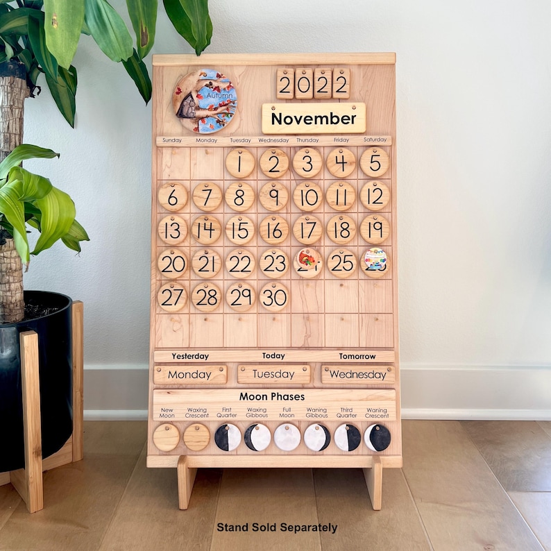 May include: A light-coloured wooden perpetual calendar board featuring the year 2022 and the month of November. It includes numbered circular wooden discs for dates, a section for days of the week, and a moon phases tracker with circular icons.