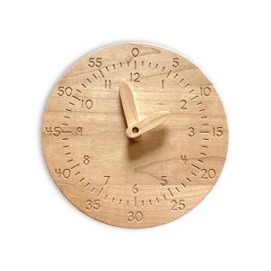Puede incluir: Un reloj redondo de madera con dos manecillas móviles. La esfera presenta un círculo interior con los números del 1 al 12 y un círculo exterior con incrementos de cinco del 0 al 55. Los números y marcas están grabados en la madera clara.