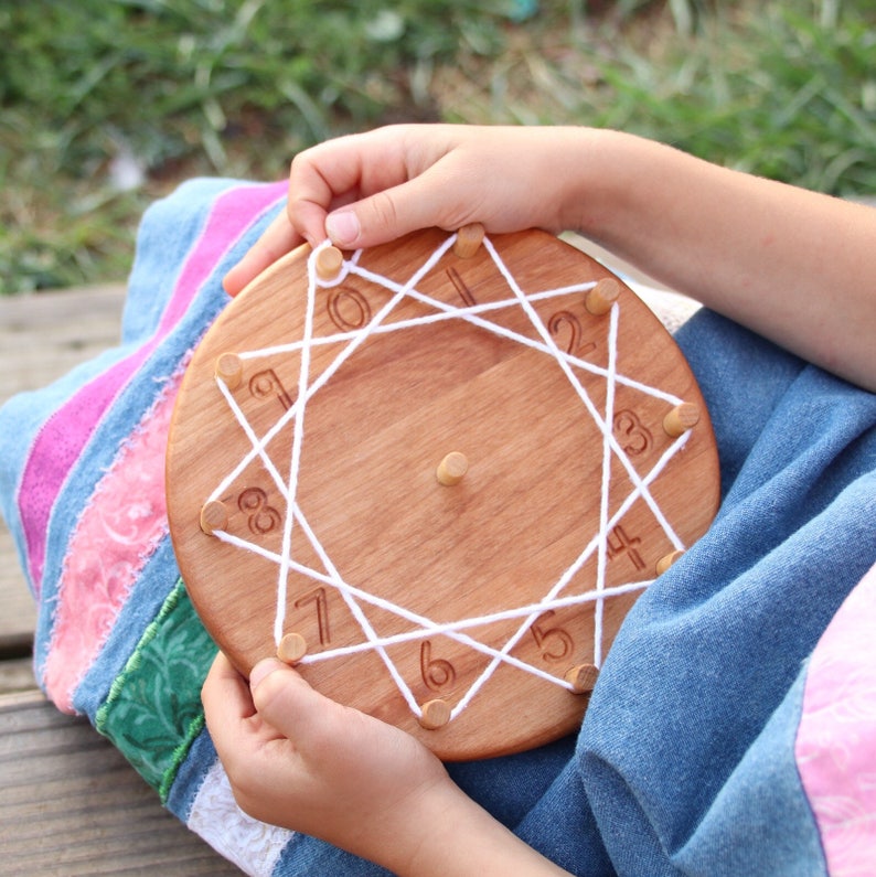 Puede incluir: Un c&iacute;rculo de madera con los n&uacute;meros del 1 al 8 alrededor del borde. Una cuerda blanca se pasa a trav&eacute;s de los agujeros del c&iacute;rculo, creando una forma de estrella. La mano de un ni&ntilde;o sostiene el c&iacute;rculo y la cuerda.