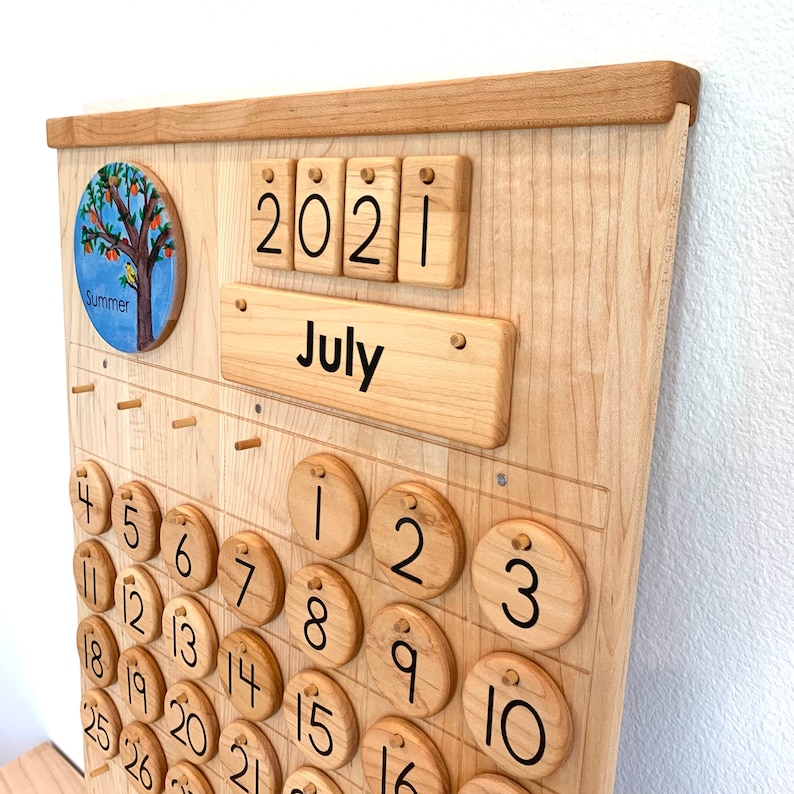 May include: A light-coloured wooden wall calendar featuring a circular summer tree illustration, a horizontal wooden plaque labelled July, and four wooden tiles displaying the year 2021. The calendar includes circular wooden number discs.