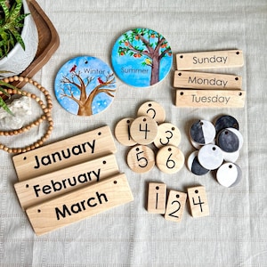 May include: A wooden perpetual calendar set featuring rectangular blocks with the months January, February, March and days Sunday, Monday, Tuesday. Includes circular discs with seasonal illustrations, numbered wooden tokens, and moon phase discs.