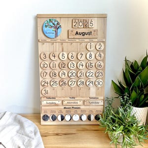 May include: A wooden perpetual calendar board featuring the year 2025 and the month of August. It includes a circular summer tree illustration, numbered wooden discs for dates 1 through 31, day-of-the-week labels, and a moon phase tracker.