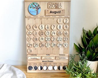 May include: A wooden perpetual calendar board featuring the year 2025 and the month of August. It includes a circular summer tree illustration, numbered wooden discs for dates 1 through 31, day-of-the-week labels, and a moon phase tracker.