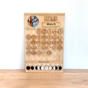May include: A wooden perpetual calendar board featuring a circular spring tree illustration, the year 2022, and the month March. It includes numbered wooden discs for dates 1 through 31 and a section for moon phases with labelled circular icons.