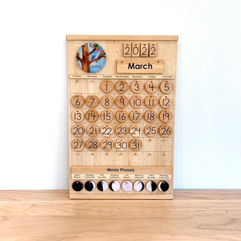 May include: A wooden perpetual calendar board featuring a circular spring tree illustration, the year 2022, and the month March. It includes numbered wooden discs for dates 1 through 31 and a section for moon phases with labelled circular icons.