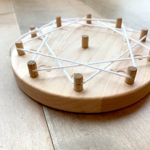 Puede incluir: Un c&iacute;rculo de madera con clavijas alrededor del borde y una cuerda blanca enhebrada entre ellas. La cuerda crea un patr&oacute;n de estrella.