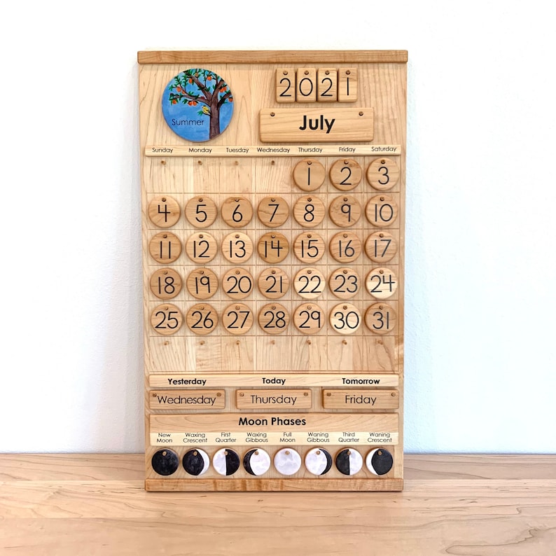 May include: A light-coloured wooden perpetual calendar board featuring a circular summer tree illustration, the year 2021, and the month of July. It includes numbered wooden discs for dates, day-of-the-week labels, and moon phase icons.