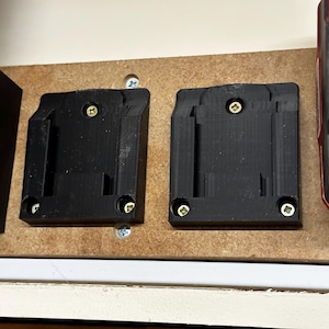 Peut inclure: Deux supports noirs en plastique pour batteries d'outils électriques, montés sur une planche de bois avec des vis dorées. Un outil électrique rouge et noir est visible à droite. Les supports sont conçus pour maintenir les batteries d'outils électriques.