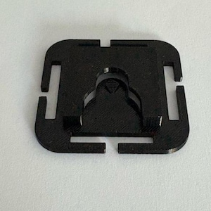 Peut inclure: Un composant en plastique noir de forme carrée, avec une découpe centrale incurvée et quatre fentes rectangulaires sur les côtés. Le composant a une finition mate et est posé sur un fond blanc.
