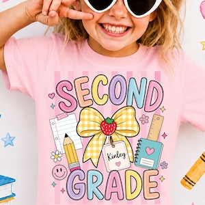 May include: A light pink t-shirt featuring a graphic print with the text SECOND GRADE in colorful bubble letters. The design includes a large yellow gingham bow with a strawberry, a pencil, a notebook, a ruler, and a tag labeled Kinley.