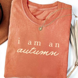 May include: A coral-colored t-shirt with the text "i am an autumn" in a cream-colored script. A gold necklace with a small pendant rests on the shirt. A brown hat and dried palm leaf are partially visible.