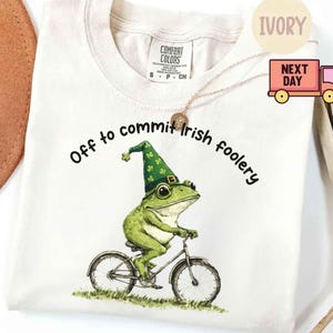 May include: Ivory-colored t-shirt with the text "Off to commit Irish foolery." A green frog wearing a green hat with shamrocks is riding a bicycle. The shirt has a Comfort Colors tag.
