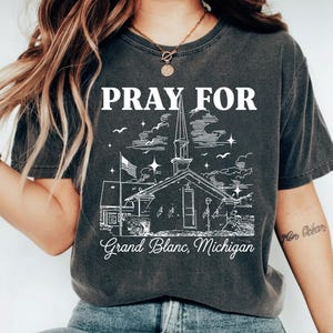 May include: Dark gray t-shirt with the words "PRAY FOR" above a white illustration of a church with an American flag. Below the church illustration is the text "Grand Blanc, Michigan".