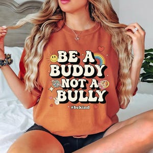 May include: An orange t-shirt with the text "BE A BUDDY NOT A BULLY" in a retro font. The design includes a smiling sun, rainbow, disco ball, peace sign, and the hashtag #bekind.
