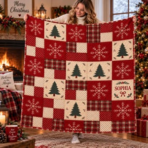 May include: A quilted throw blanket featuring a patchwork design of red, cream, and green squares. The pattern includes white snowflakes, dark green Christmas trees, and red plaid squares. The text SOPHIA is printed in red with a small bow icon.