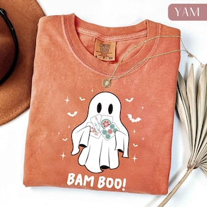 May include: A burnt orange Comfort Colors t-shirt featuring a white ghost graphic holding playing cards. The shirt has the text "BAM BOO!" and is accessorized with a gold necklace. Bats and stars are also printed on the shirt.