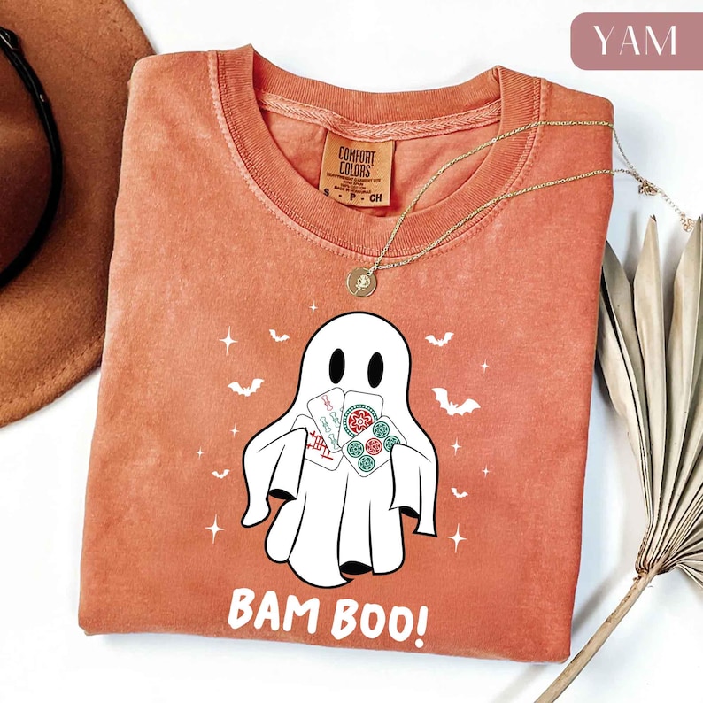 May include: A burnt orange Comfort Colors t-shirt featuring a white ghost graphic holding playing cards. The shirt has the text "BAM BOO!" and is accessorized with a gold necklace. Bats and stars are also printed on the shirt.