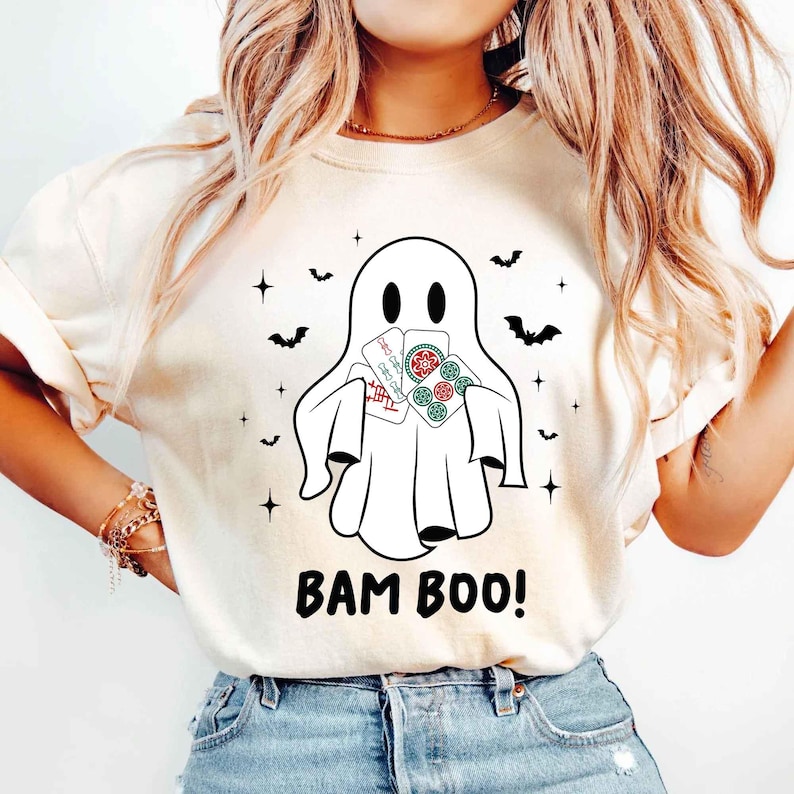 May include: A cream-coloured t-shirt with a cartoon ghost holding playing cards. The ghost has a simple design with two eye holes and a wide mouth. The text "BAM BOO!" is printed in black below the ghost, with bats and stars around the ghost.