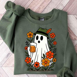 May include: Olive green sweatshirt featuring a cartoon ghost wearing a green hat with a feather, holding a beer stein. The ghost is surrounded by orange and yellow flowers and green foliage. The design is centered on the front of the sweatshirt.