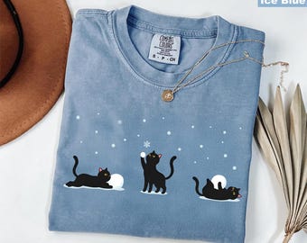 May include: Ice blue t-shirt with a crew neck and short sleeves. The shirt features a graphic of three black cats playing in the snow. The cats are depicted with yellow eyes and pink tongues. The shirt has a Comfort Colors tag.
