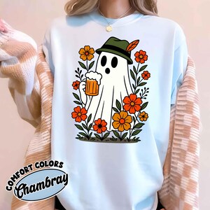 May include: Light blue sweatshirt with a graphic of a cartoon ghost wearing a green hat and holding a beer stein. The ghost is surrounded by orange and yellow flowers and green leaves. The text "COMFORT COLORS Chambray" is printed on the bottom left.