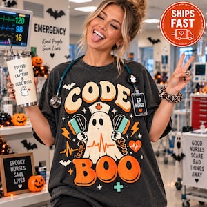 May include: A charcoal gray t-shirt featuring a graphic of a ghost wearing a nurse cap and holding defibrillator paddles. The text reads "CODE BOO" with an orange heart and heartbeat line. A white tumbler with the text "FUELED BY CAFFEINE & CODE BOOS" is held in one hand.