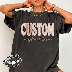 May include: A dark grey t-shirt with the word "CUSTOM" in a large, decorative font. The letters have a pink and white pattern. Below the word "CUSTOM" is the phrase "optional line" in a cursive font. The shirt also has the words "COMFORT COLORS Pepper" in a circular design.