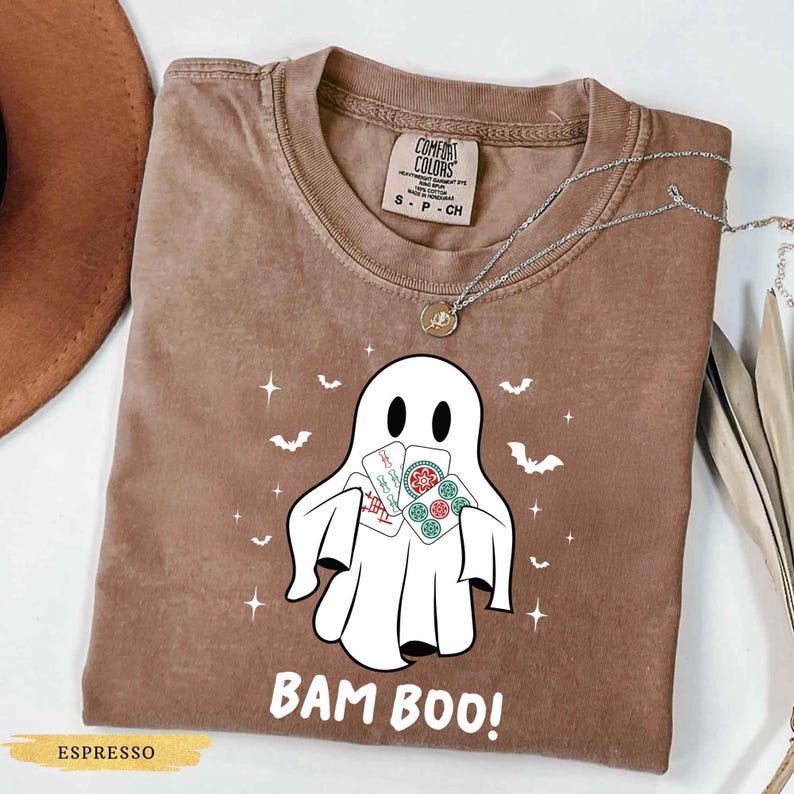 May include: Espresso-coloured t-shirt with a white ghost graphic holding cards. The shirt features the text "BAM BOO!" in white, with small bat and star illustrations. A silver necklace with a pendant is also visible.