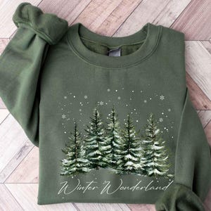 May include: An olive green sweatshirt featuring a winter scene with snow-covered pine trees and falling snowflakes. The words "Winter Wonderland" are written in a cursive font across the bottom of the design. The sweatshirt has long sleeves.