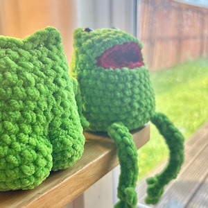May include: Two handmade green crochet frog plushies. One frog is sitting with its legs dangling, and the other is a close-up of the frog's back. The frog has a red mouth and black eyes. The plushies are on a wooden surface.
