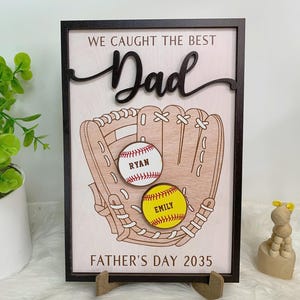 Puede incluir: Letrero de madera para el Día del Padre con un diseño de guante de béisbol. El letrero dice "WE CAUGHT THE BEST Dad" y "FATHER'S DAY 2035". Dos pelotas de béisbol están en el guante, una con "RYAN" y la otra con "EMILY". El letrero está enmarcado en negro.