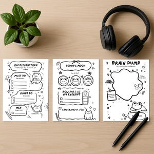 May include: Three black and white printable planner pages with frog illustrations and text. The pages include sections for to-do lists, mood tracking, and a brain dump. A pair of black pens, a potted plant, and black headphones are also in the image.