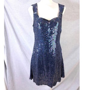 May include: A sleeveless knee-length dress covered in dark blue sequins. The garment features a square neckline and wide shoulder straps. The sequins are arranged in vertical lines, with a concentrated cluster of iridescent light blue accents.