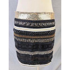 May include: A black mini skirt with multiple embellished bands. The top band is gold sequins, followed by bands of silver, black, and gold beads, and a white beaded trim at the bottom. The skirt is displayed on a mannequin.